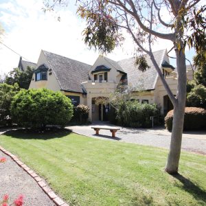 A double storey, yellow building in a garden with a green lawn and gum tree. It's sunny. 